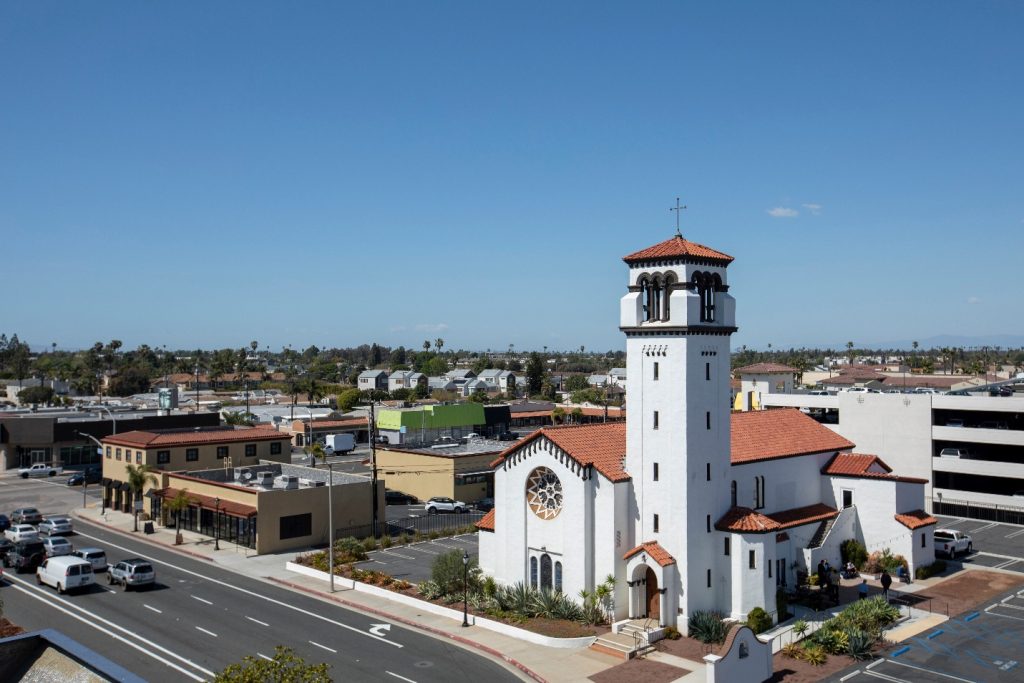 White church with red-tile roof and bell tower in Costa Mesa, CA, with parked cars, palm trees, and suburban skyline