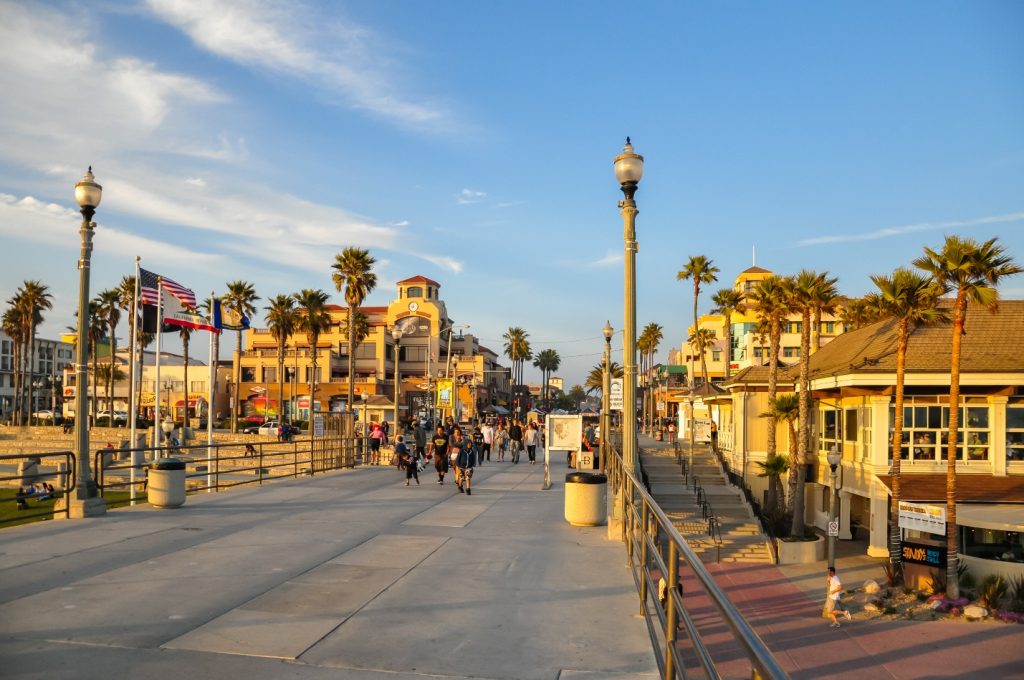 Busy boardwalk in Huntington Beach, CA with palm trees, shops, flags, and people walking under a clear blue sky at sunset