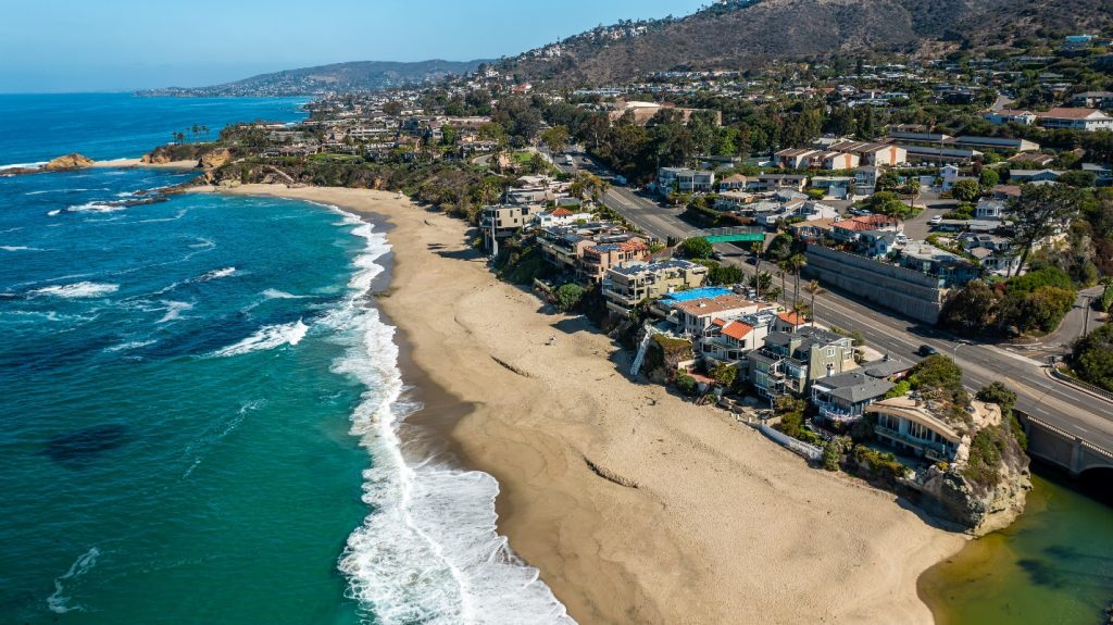 Aerial view of Laguna Beach, CA featuring ocean waves, sandy shoreline, coastal homes, and scenic hills along Pacific Coast Highway