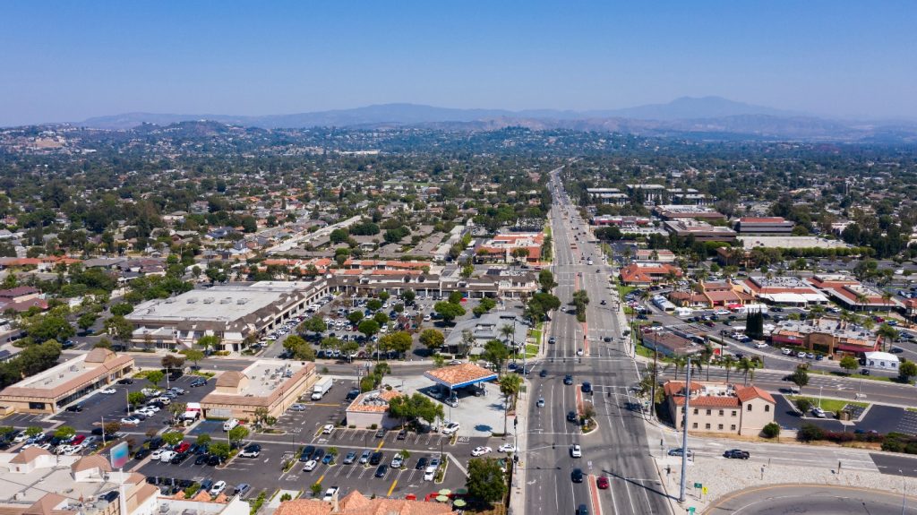 Aerial view of Tustin, CA featuring a major road lined with cars, commercial buildings, residential areas, and distant mountains