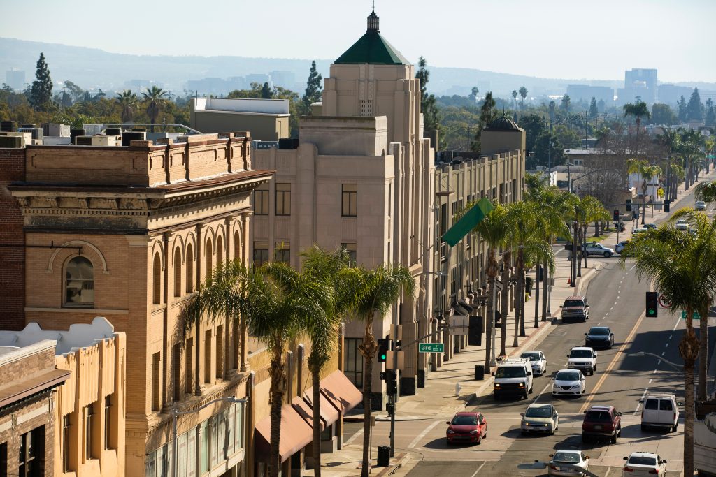 Street view of downtown area with historic brick buildings, palm trees lining the sidewalks, and cars driving along a busy road in the afternoon sunlight