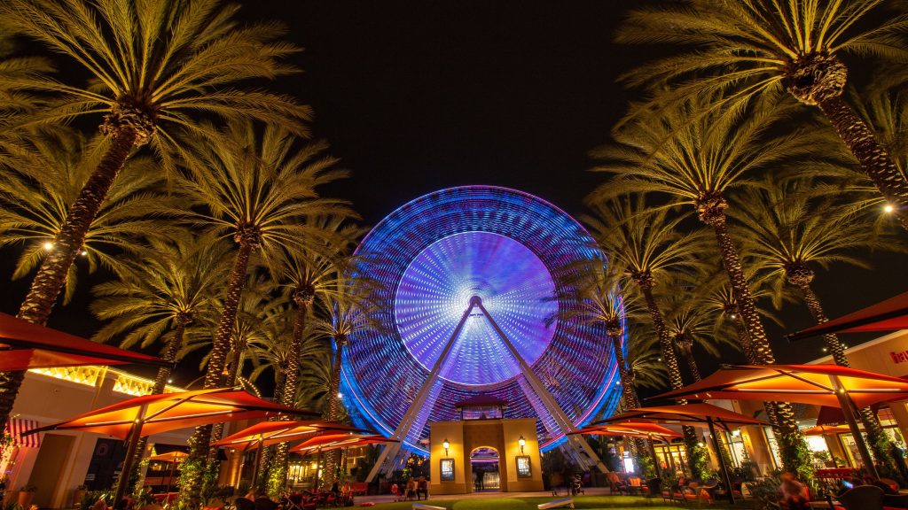 Colorful Ferris wheel illuminated at night surrounded by tall palm trees and outdoor dining umbrellas at Irvine Spectrum Center in California