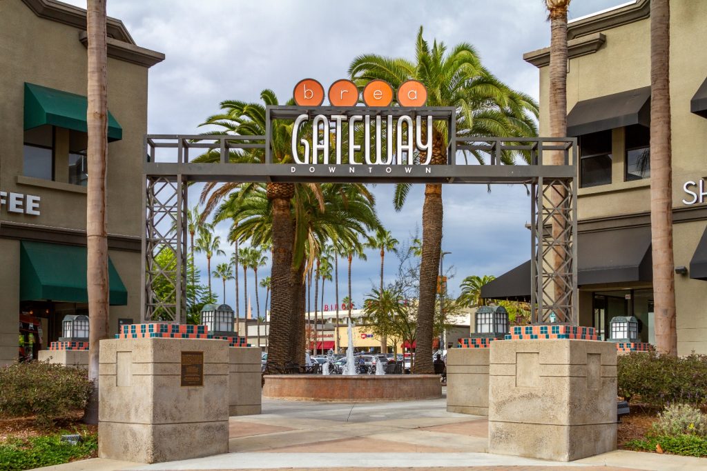 Entrance to Downtown Brea Gateway in Brea, California, featuring a modern metal archway, palm trees, shops, and a fountain under a cloudy sky.