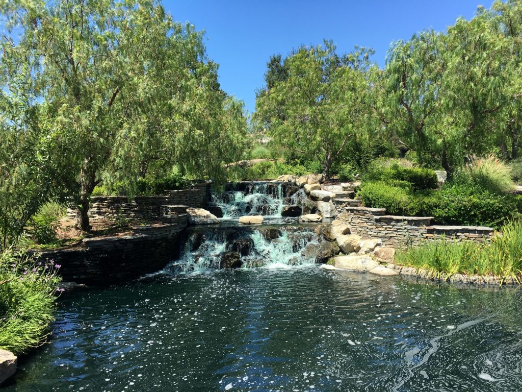 Scenic view of a waterfall and pond surrounded by lush greenery and stone landscaping in Coto de Caza, California, under a clear blue sky.
