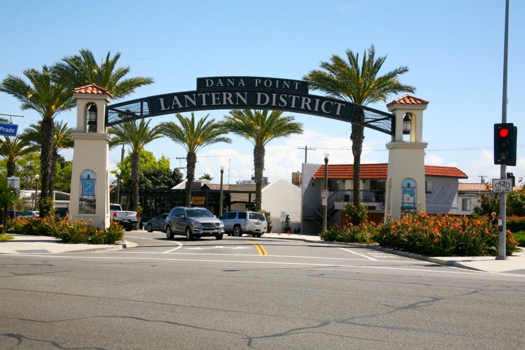 Entrance to the Lantern District in Dana Point, California, featuring palm trees, a large black archway sign, and cars driving through a lively downtown area under a clear blue sky.