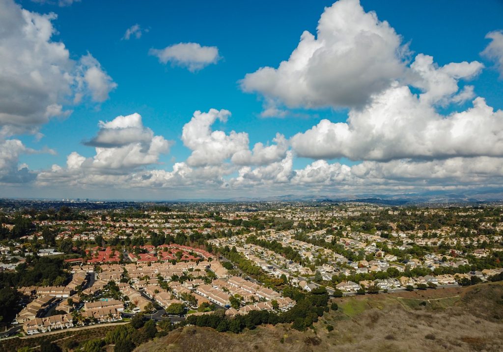 Aerial view of suburban neighborhoods in Ladera Ranch, California, with clusters of homes, green hills, and large white clouds under a bright blue sky.