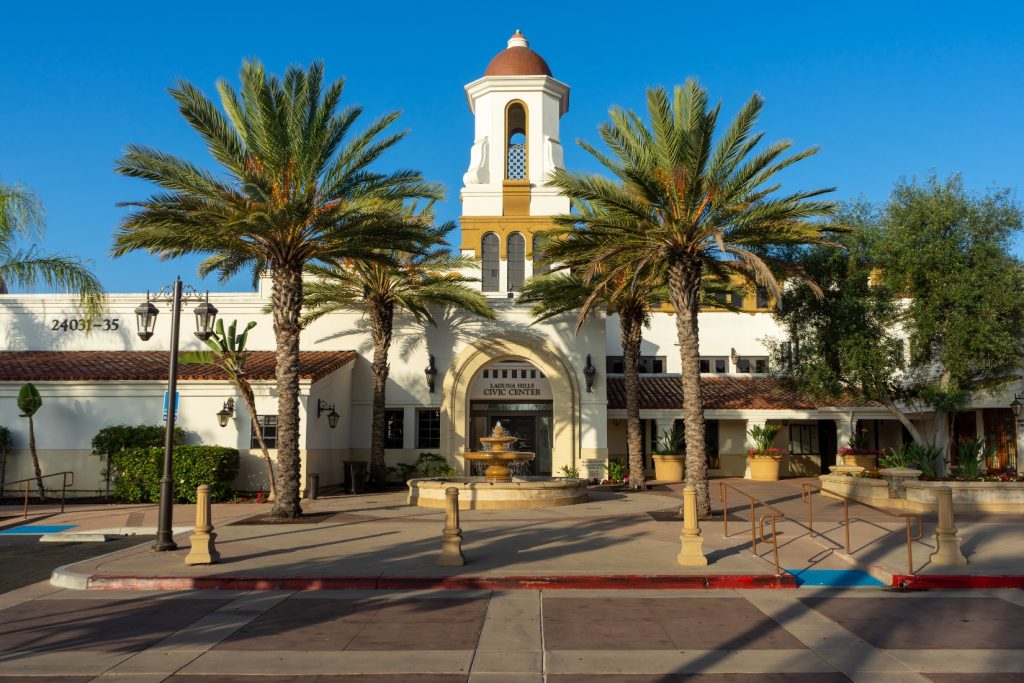 Front view of Laguna Hills Civic Center in Laguna Hills, California, featuring palm trees, a fountain, and Spanish-style architecture under a clear blue sky.