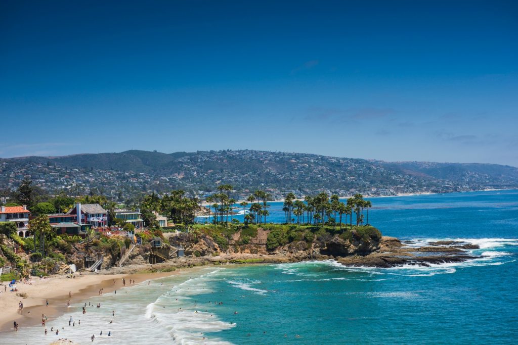 Scenic coastal view near Laguna Niguel, California, showing sandy beaches, rocky cliffs, palm trees, and clear blue ocean waters under a bright sky.