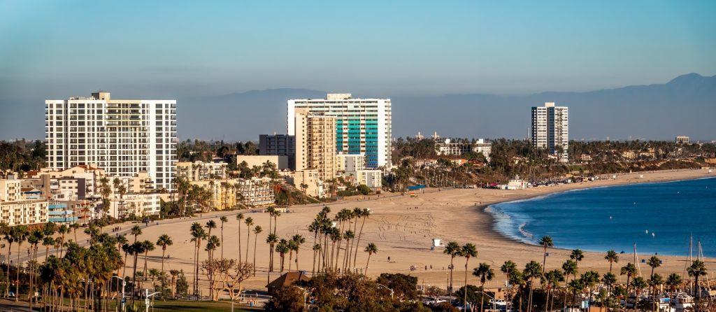 View of Los Alamitos, California coastline with high-rise buildings, sandy beaches, palm trees, and the ocean under a clear blue sky.