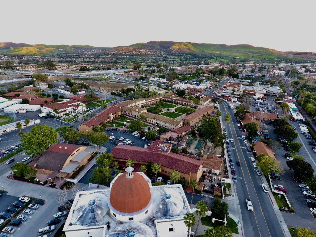 Aerial view of historic San Juan Capistrano, California, featuring the Mission San Juan Capistrano, surrounding streets, and green hills in the background.
